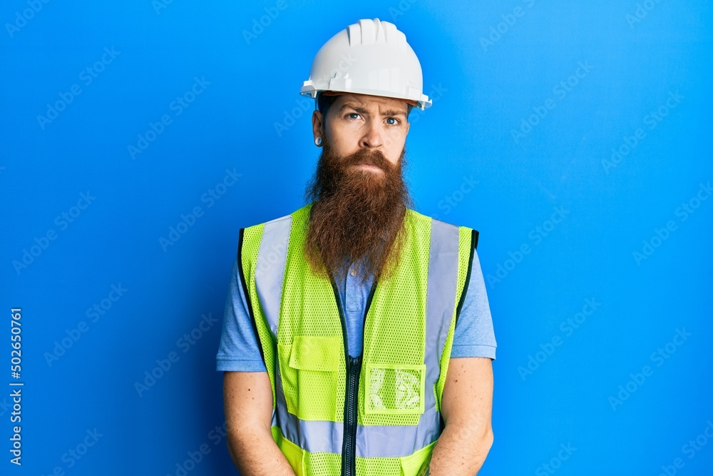 Redhead man with long beard wearing safety helmet and reflective jacket relaxed with serious expression on face. simple and natural looking at the camera. Redhead man with long beard wearing safety helmet and reflective jacket relaxed with serious expression on face. simple and natural looking at the camera.