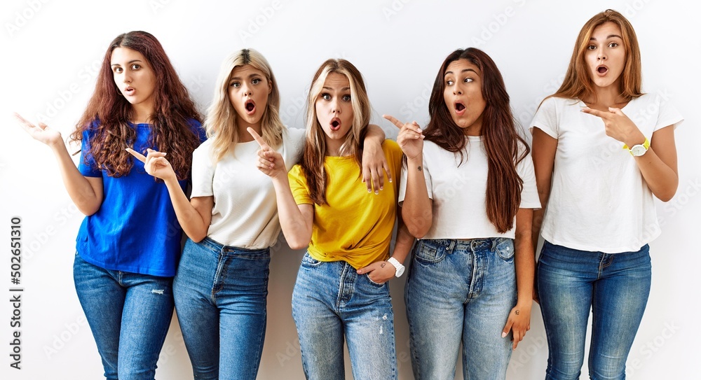 Group of young girl friends standing together over isolated background surprised pointing with finger to the side, open mouth amazed expression. Group of young girl friends standing together over isolated background surprised pointing with finger to the side, open mouth amazed expression.