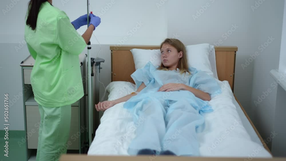 Wide shot portrait of hopeless teenage girl lying in bed in ward with nurse putting dropper to patient. Sad frustrated Caucasian teenager with fatal illness undergoing treatment in hospital Wide shot portrait of hopeless teenage girl lying in bed in ward with nurse putting dropper to patient. Sad frustrated Caucasian teenager with fatal illness undergoing treatment in hospital
