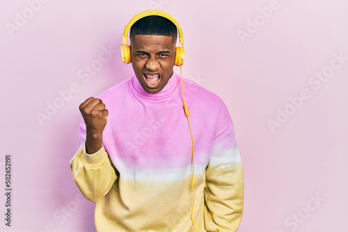 Young black man listening to music wearing headphones angry and mad raising fist frustrated and furious while shouting with anger. rage and aggressive concept.