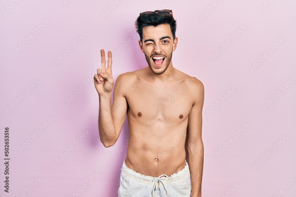 Young hispanic man wearing swimwear shirtless smiling with happy face winking at the camera doing victory sign. number two. Young hispanic man wearing swimwear shirtless smiling with happy face winking at the camera doing victory sign. number two.