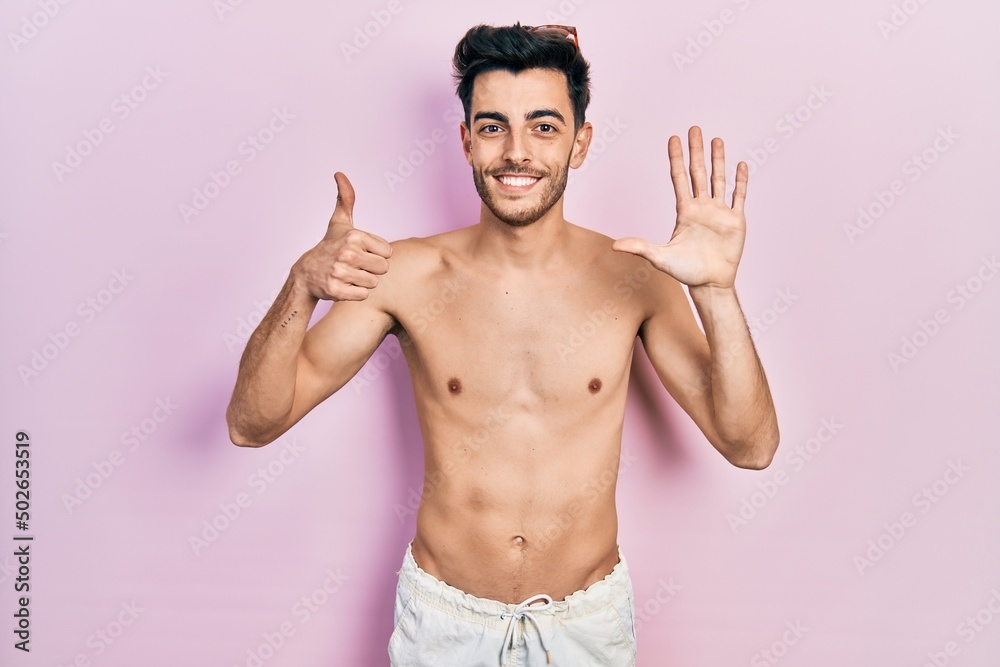 Young hispanic man wearing swimwear shirtless showing and pointing up with fingers number six while smiling confident and happy.