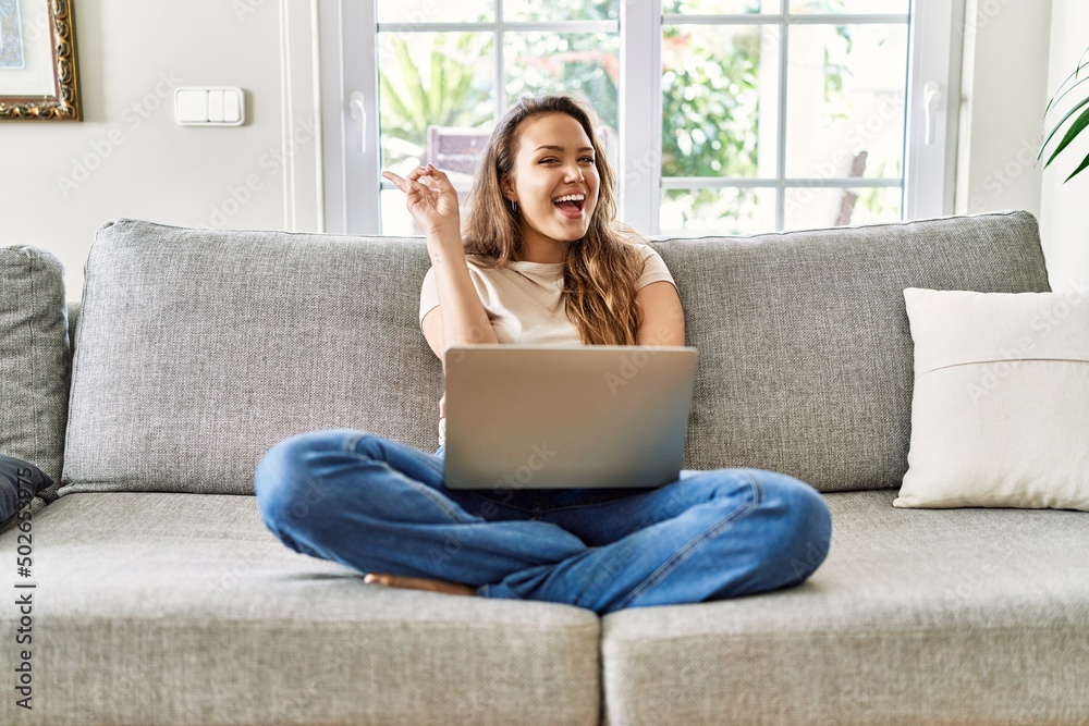 Beautiful young brunette woman sitting on the sofa using computer laptop at home smiling with happy face winking at the camera doing victory sign. number two. Beautiful young brunette woman sitting on the sofa using computer laptop at home smiling with happy face winking at the camera doing victory sign. number two.