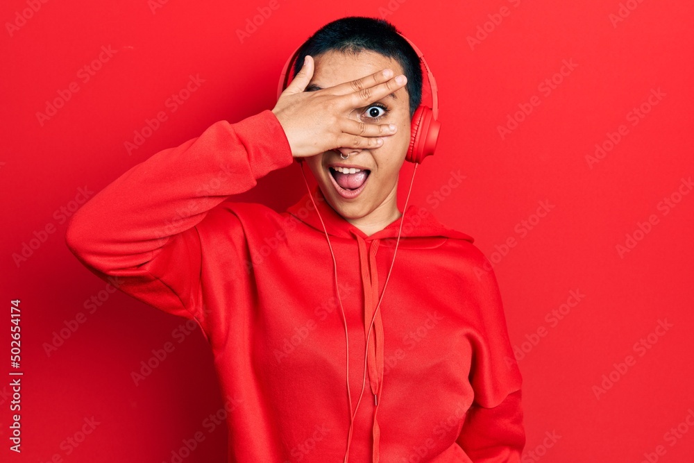 Naklejka premium Beautiful hispanic woman with short hair listening to music using headphones peeking in shock covering face and eyes with hand, looking through fingers with embarrassed expression.