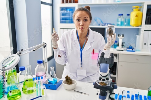 Young hispanic woman doing weed oil extraction at laboratory making fish face with mouth and squinting eyes, crazy and comical.