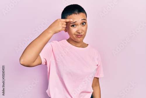 Beautiful hispanic woman with short hair wearing casual pink t shirt pointing unhappy to pimple on forehead, ugly infection of blackhead. acne and skin problem