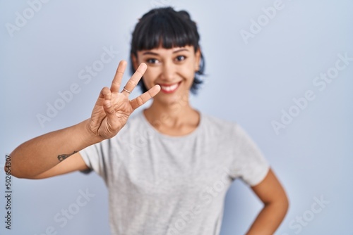 Young hispanic woman wearing casual t shirt over blue background showing and pointing up with fingers number three while smiling confident and happy.