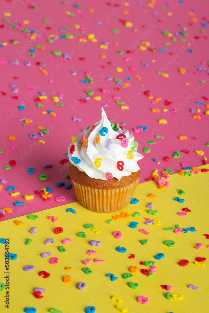 Cupcakes with white frosting and assorted sprinkles placed on a pink and yellow surface.