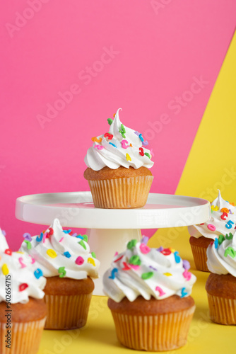 Cupcakes with creamy topping and sprinkles displayed on a tiered stand  contrasting pink and yellow backdrop.