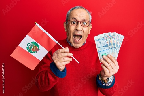 Handsome senior man with grey hair holding peru flag and peruvian sol banknotes celebrating crazy and amazed for success with open eyes screaming excited. photo
