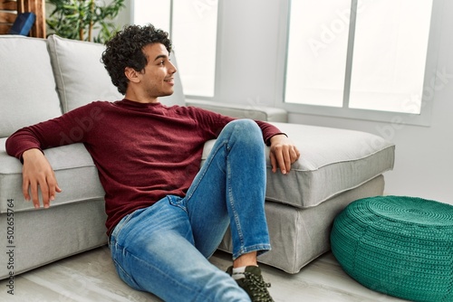 Young hispanic man smiling happy sitting on the floor at home.