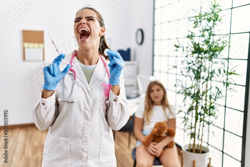 Young doctor woman putting vaccine to little girl angry and mad screaming frustrated and furious, shouting with anger looking up.