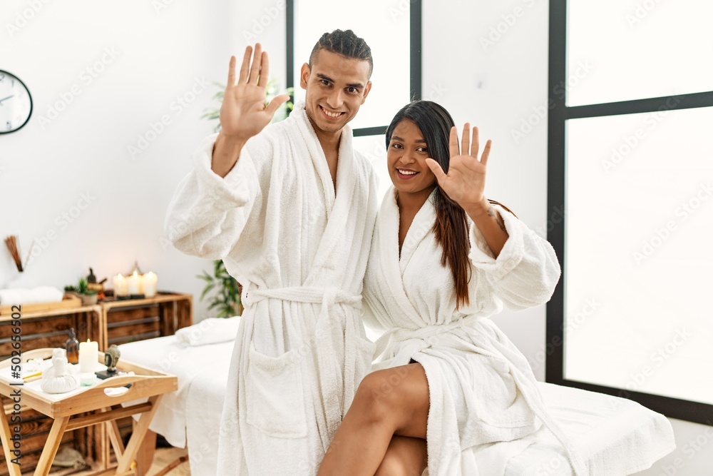 Young latin couple wearing towel standing at beauty center waiving saying hello happy and smiling, friendly welcome gesture Young latin couple wearing towel standing at beauty center waiving saying hello happy and smiling, friendly welcome gesture