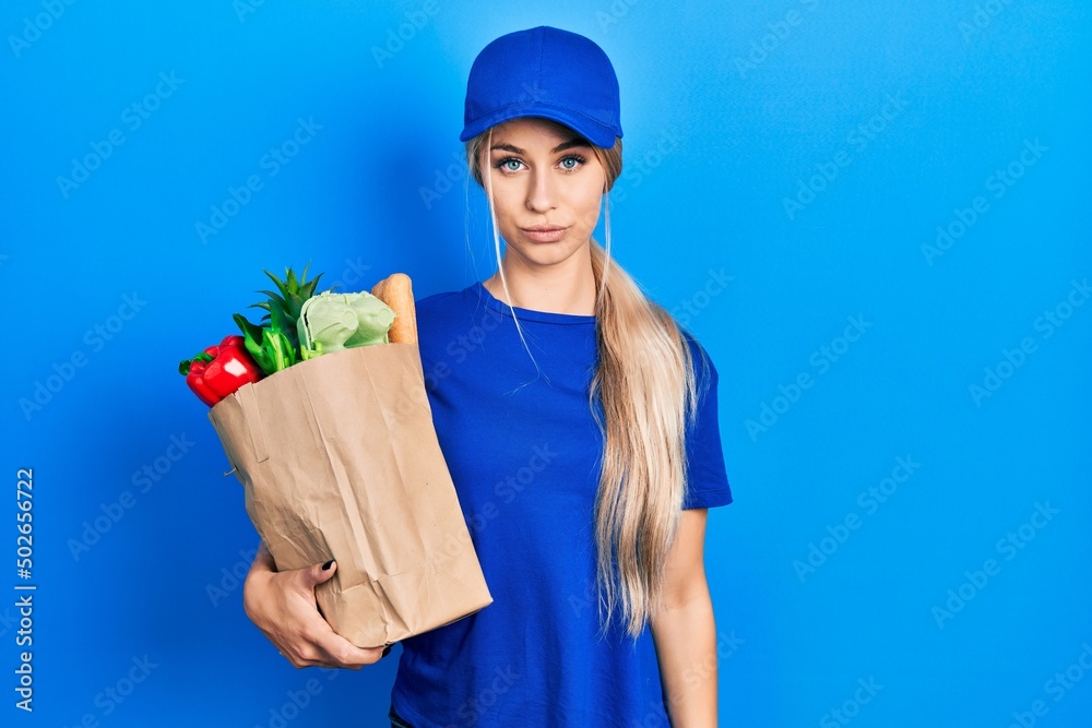 Young caucasian woman wearing courier uniform with groceries from supermarket relaxed with serious expression on face. simple and natural looking at the camera. Young caucasian woman wearing courier uniform with groceries from supermarket relaxed with serious expression on face. simple and natural looking at the camera.