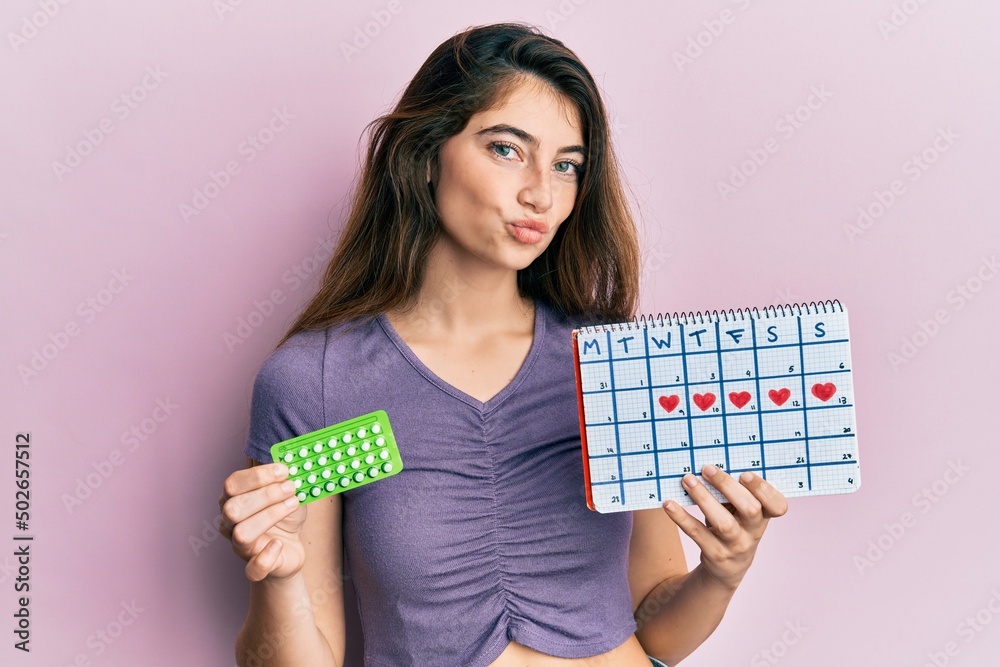 Young caucasian woman holding birth control pills and menstrual calendar looking at the camera blowing a kiss being lovely and sexy. love expression. Young caucasian woman holding birth control pills and menstrual calendar looking at the camera blowing a kiss being lovely and sexy. love expression.