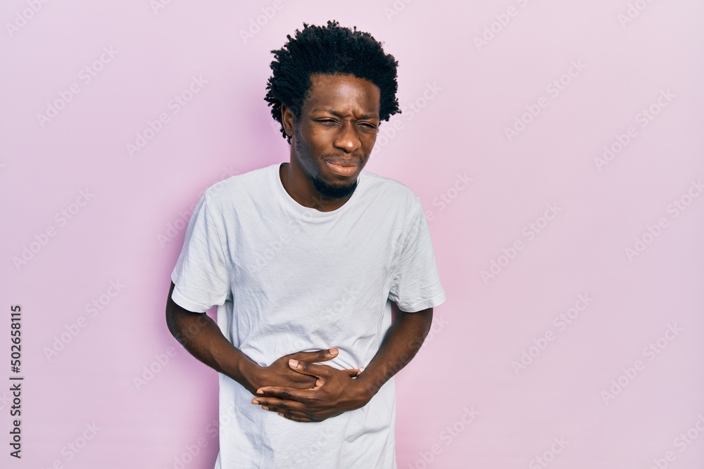Young african american man wearing casual white t shirt with hand on stomach because indigestion, painful illness feeling unwell. ache concept. Young african american man wearing casual white t shirt with hand on stomach because indigestion, painful illness feeling unwell. ache concept.