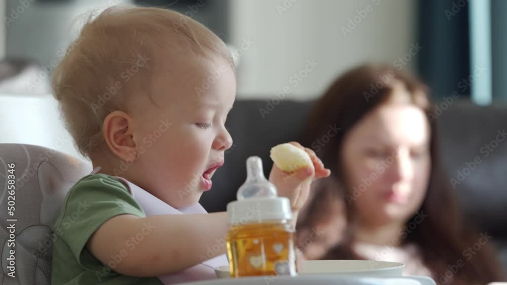 Baby boy with mother in the living room, cute kid sitting in booster seat and eating an apple, mom using smartphone. High quality 4k footage Baby boy with mother in the living room, cute kid sitting in booster seat and eating an apple, mom using smartphone. High quality 4k footage