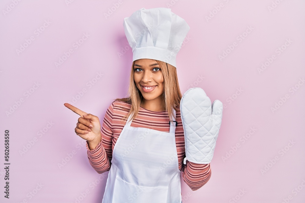 Beautiful hispanic woman uniform wearing apron and hat wearing protective glove smiling happy pointing with hand and finger to the side