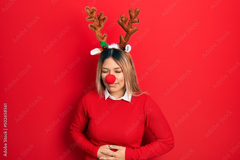 Beautiful hispanic woman wearing deer christmas hat and red nose with hand on stomach because indigestion, painful illness feeling unwell. ache concept. Beautiful hispanic woman wearing deer christmas hat and red nose with hand on stomach because indigestion, painful illness feeling unwell. ache concept.