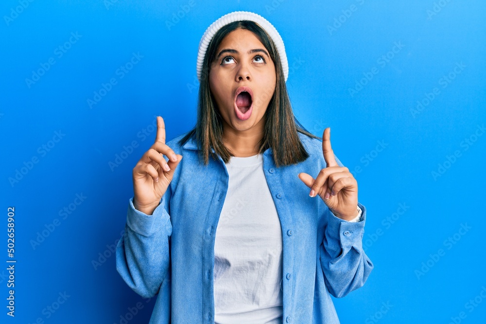 Young latin woman wearing cute wool cap amazed and surprised looking up and pointing with fingers and raised arms. Young latin woman wearing cute wool cap amazed and surprised looking up and pointing with fingers and raised arms.
