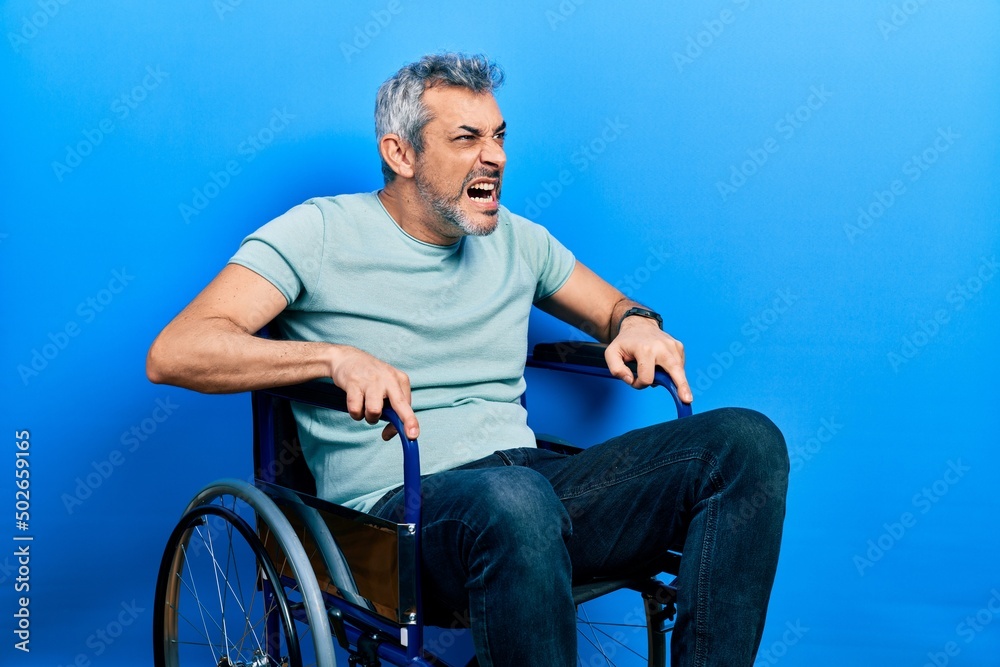 Fototapeta premium Handsome middle age man with grey hair sitting on wheelchair angry and mad screaming frustrated and furious, shouting with anger. rage and aggressive concept.