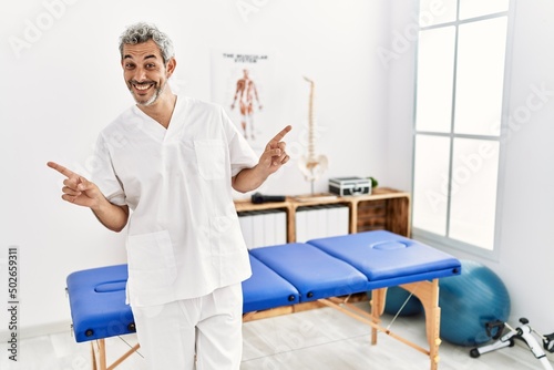 Middle age hispanic therapist man working at pain recovery clinic smiling confident pointing with fingers to different directions. copy space for advertisement