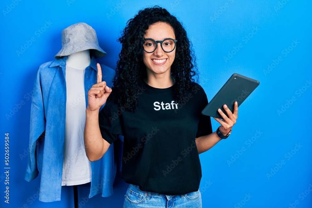Young hispanic woman with curly hair wearing staff t shirt holding touchpad device smiling with an idea or question pointing finger with happy face, number one Young hispanic woman with curly hair wearing staff t shirt holding touchpad device smiling with an idea or question pointing finger with happy face, number one