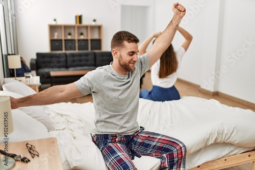 Young caucasian couple yawning and stretching arms sitting on the bed.