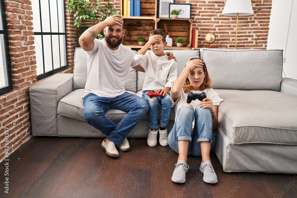 Family of three playing video game sitting on the sofa stressed and frustrated with hand on head, surprised and angry face