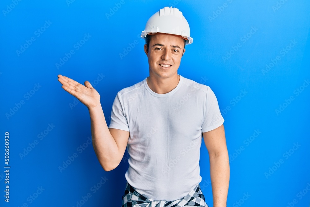 Handsome young man wearing builder uniform and hardhat smiling cheerful presenting and pointing with palm of hand looking at the camera. Handsome young man wearing builder uniform and hardhat smiling cheerful presenting and pointing with palm of hand looking at the camera.