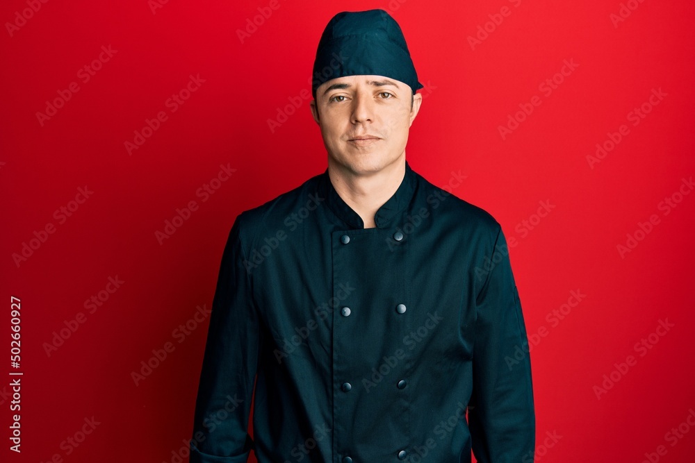 Handsome young man wearing professional cook uniform and hat relaxed with serious expression on face. simple and natural looking at the camera. Handsome young man wearing professional cook uniform and hat relaxed with serious expression on face. simple and natural looking at the camera.