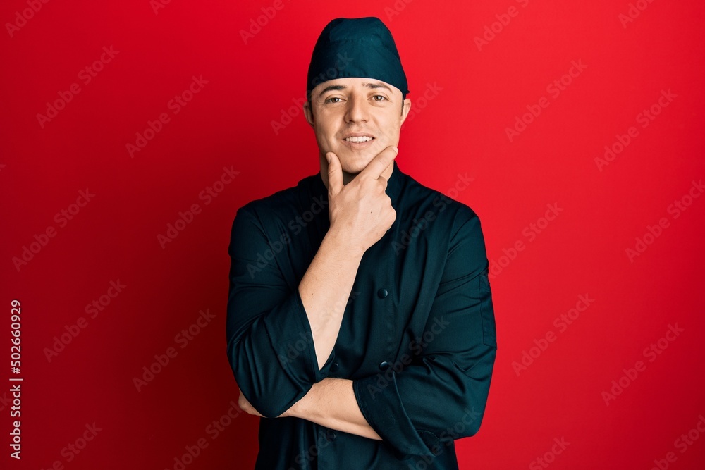 Handsome young man wearing professional cook uniform and hat looking confident at the camera smiling with crossed arms and hand raised on chin. thinking positive. Handsome young man wearing professional cook uniform and hat looking confident at the camera smiling with crossed arms and hand raised on chin. thinking positive.