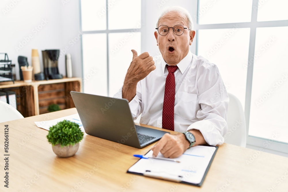 Senior man working at the office using computer laptop surprised pointing with hand finger to the side, open mouth amazed expression. Senior man working at the office using computer laptop surprised pointing with hand finger to the side, open mouth amazed expression.