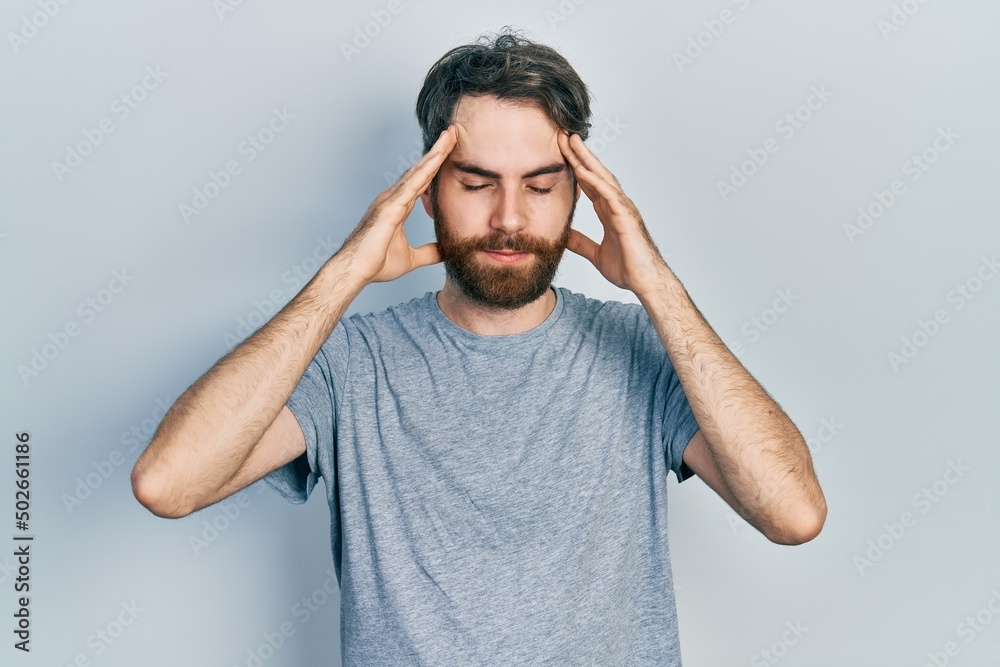 Caucasian man with beard wearing casual grey t shirt with hand on head for pain in head because stress. suffering migraine. Caucasian man with beard wearing casual grey t shirt with hand on head for pain in head because stress. suffering migraine.