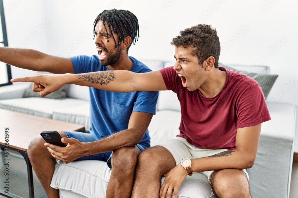 Young hispanic men using smartphone sitting on the sofa at home pointing displeased and frustrated to the camera, angry and furious with you