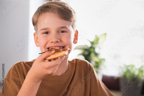 Satisfied boy enjoys eating delicious pizza in the kitchen at home.