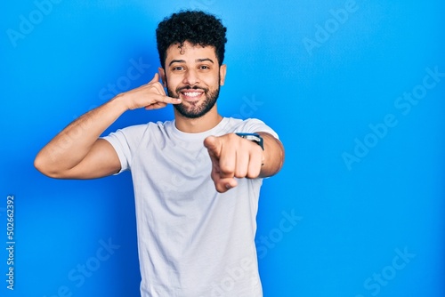 Young arab man with beard wearing casual white t shirt smiling doing talking on the telephone gesture and pointing to you. call me.