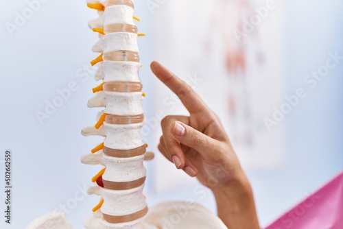 Young hispanic woman wearing physiotherapist uniform pointing to anatomical model of vertebral column at physiotherpy clinic photo