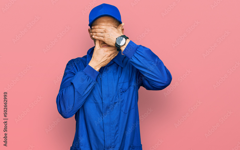 Bald man with beard wearing builder jumpsuit uniform covering eyes and mouth with hands, surprised and shocked. hiding emotion Bald man with beard wearing builder jumpsuit uniform covering eyes and mouth with hands, surprised and shocked. hiding emotion