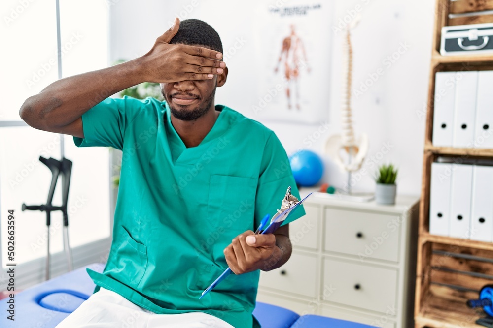 Obraz premium Young african american man working at pain recovery clinic covering eyes with hand, looking serious and sad. sightless, hiding and rejection concept