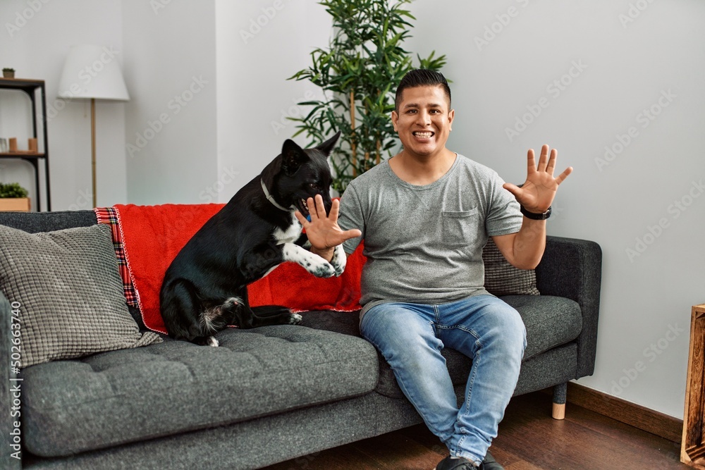 Young latin man and dog sitting on the sofa at home showing and pointing up with fingers number ten while smiling confident and happy. Young latin man and dog sitting on the sofa at home showing and pointing up with fingers number ten while smiling confident and happy.