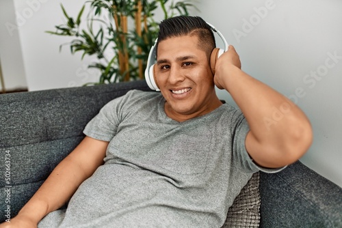 Young hispanic man smiling happy listening music sitting on the sofa at home. © Krakenimages.com