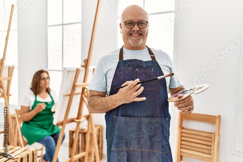Middle age hispanic man at art studio looking positive and happy standing and smiling with a confident smile showing teeth