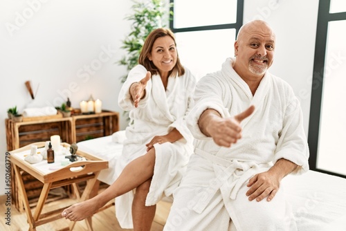 Middle age hispanic couple wearing bathrobe at wellness spa smiling friendly offering handshake as greeting and welcoming. successful business.