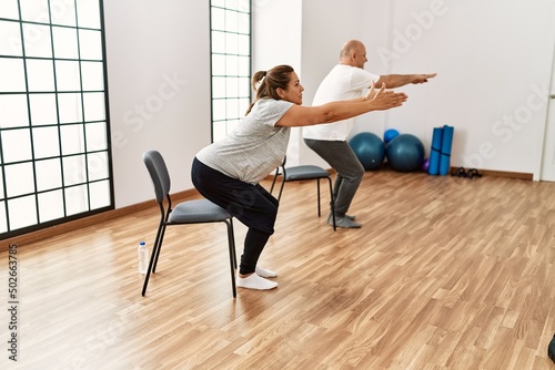 Middle age hispanic couple stretching using chair at sport center.