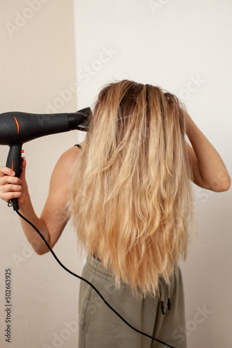 a girl with blond hair dries them with a hair dryer on a light background