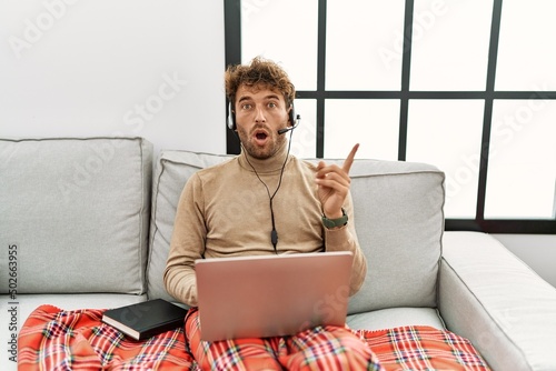 Young handsome man with beard wearing operator headset working from home surprised pointing with finger to the side, open mouth amazed expression.