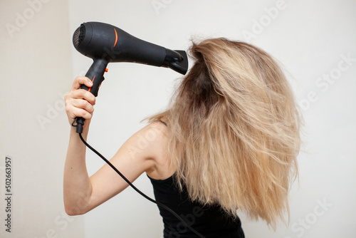 a girl with blond hair dries them with a hair dryer on a light background