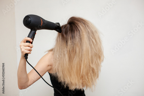 a girl with blond hair dries them with a hair dryer on a light background