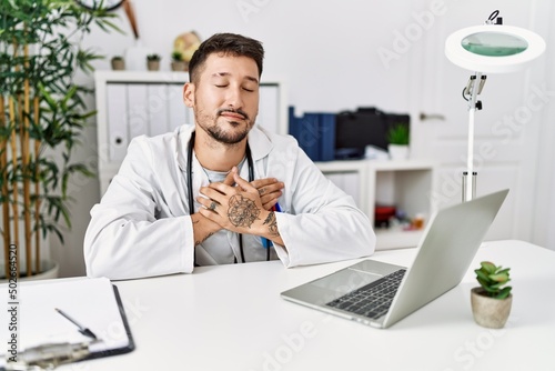 Young doctor working at the clinic using computer laptop smiling with hands on chest with closed eyes and grateful gesture on face. health concept.